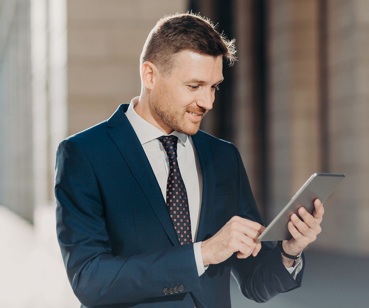 Horizontal shot of successful male office worker dressed in elegant suit, holds modern digital tablet, updates website with financial or business news. Happy trader with modern electronic device